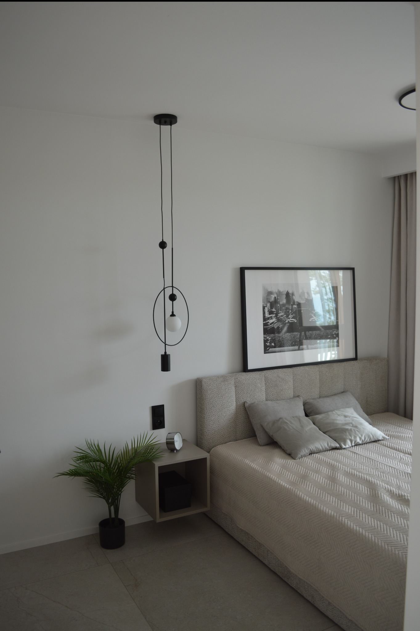 Nowoczesna sypialnia w Kołobrzegu: beżowe łóżko z szarą pościelą, minimalistyczna lampa wisząca i czarno-biały obraz nad wezgłowiem. Stonowana kolorystyka, elegancki design.