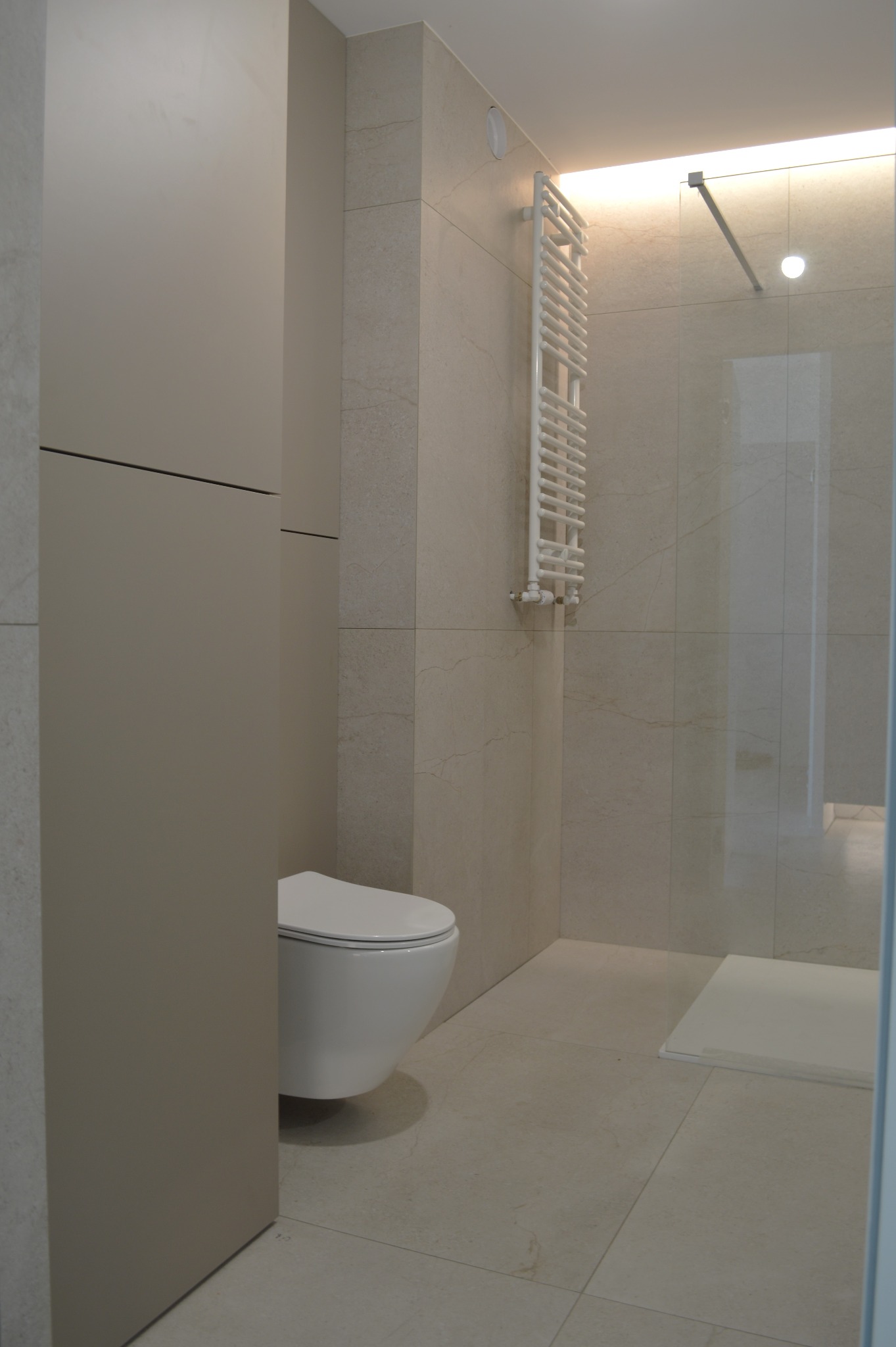 Nowoczesna łazienka w beżowych płytkach z podwieszaną toaletą, grzejnikiem drabinkowym i kabiną prysznicową. Minimalistyczny design z subtelnym oświetleniem.