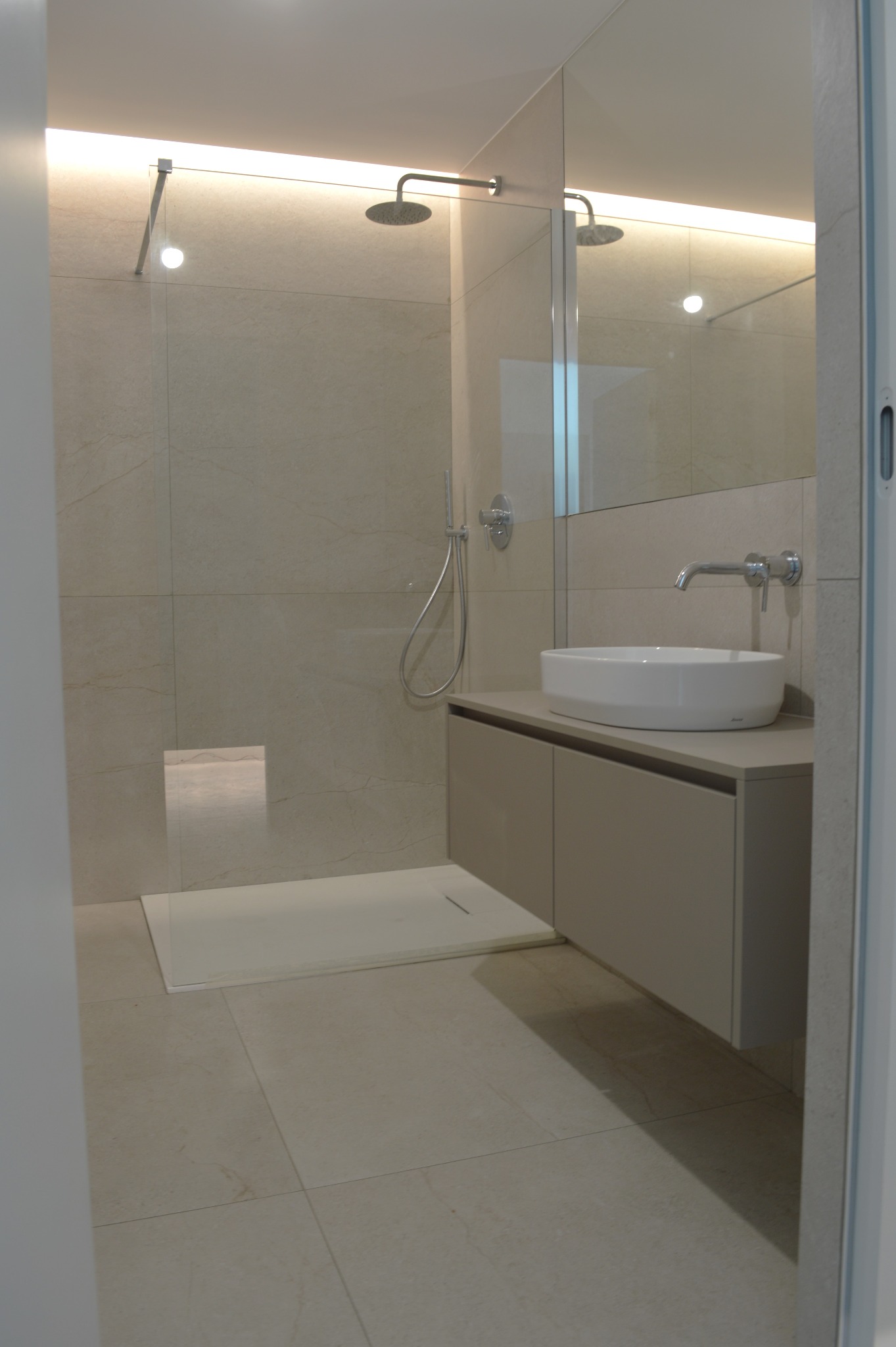 Nowoczesna łazienka z beżowymi płytkami, kabiną prysznicową typu walk-in i szafką pod umywalką z owalną białą umywalką. Minimalistyczny design, oświetlenie LED.