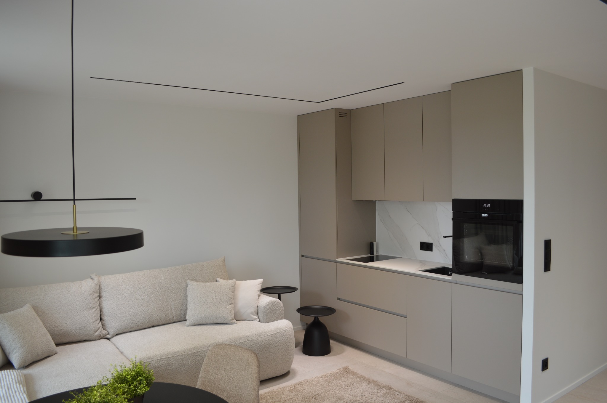 Nowoczesne wnętrze: beżowa zabudowa kuchenna z piekarnikiem, marmurowy backsplash, minimalistyczne oświetlenie i sofa z poduszkami w salonie.