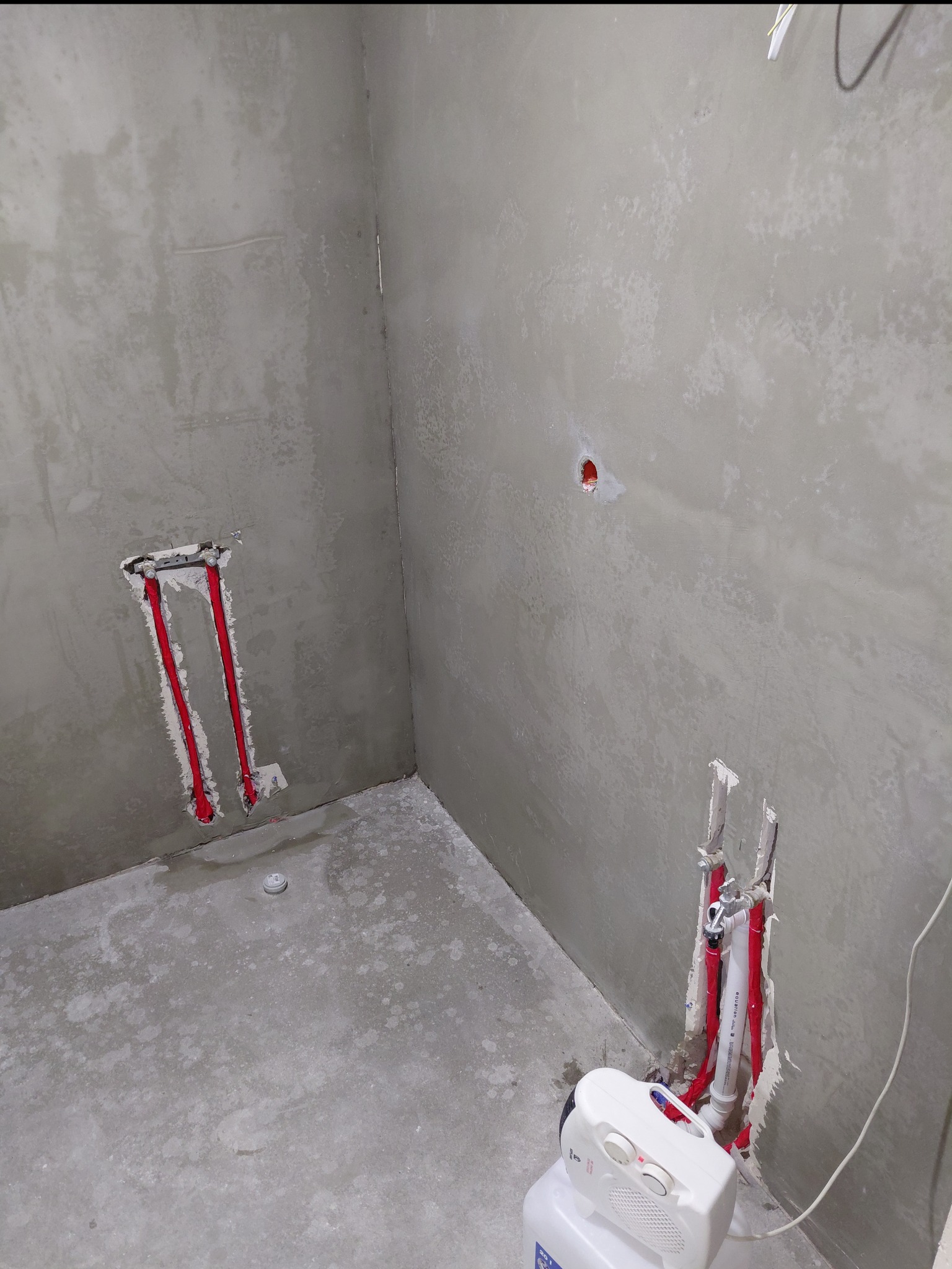 Surowa łazienka w trakcie prac: widoczne instalacje wodne w ścianach, przygotowane do dalszych etapów. Szare ściany i podłoga, brak wykończenia.