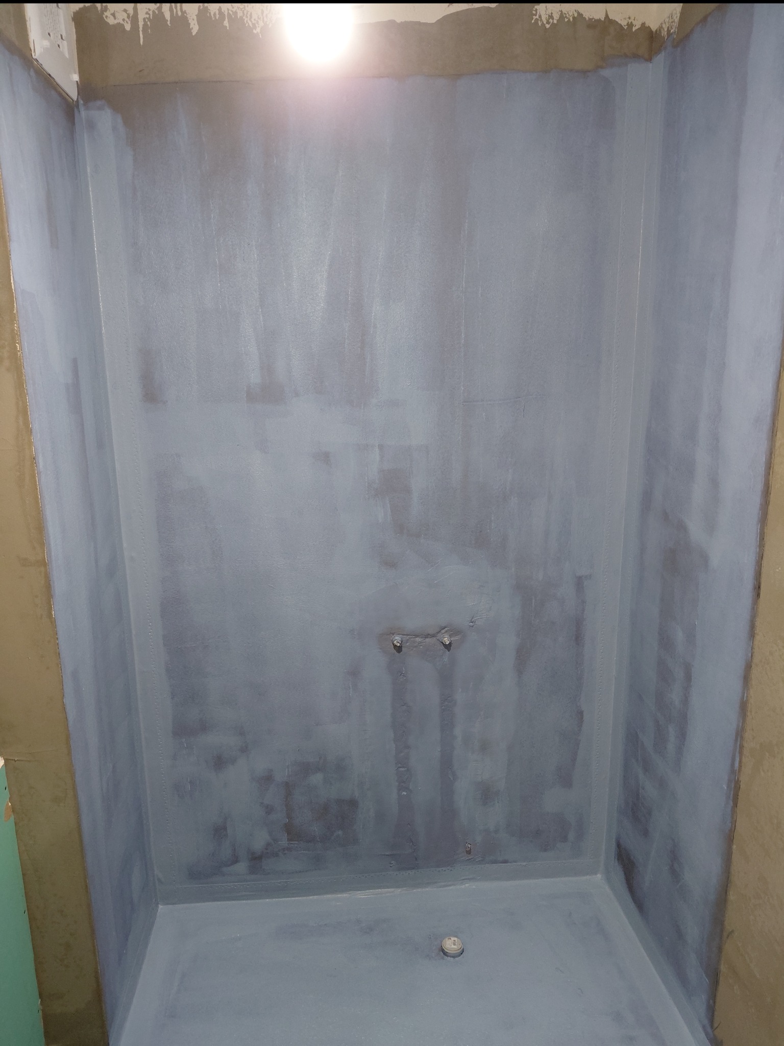 Hydroizolacja w rogu łazienki, zabezpieczająca przed wilgocią. Szare ściany i podłoga, widoczne łączenia taśm uszczelniających. Przygotowanie do dalszych prac.