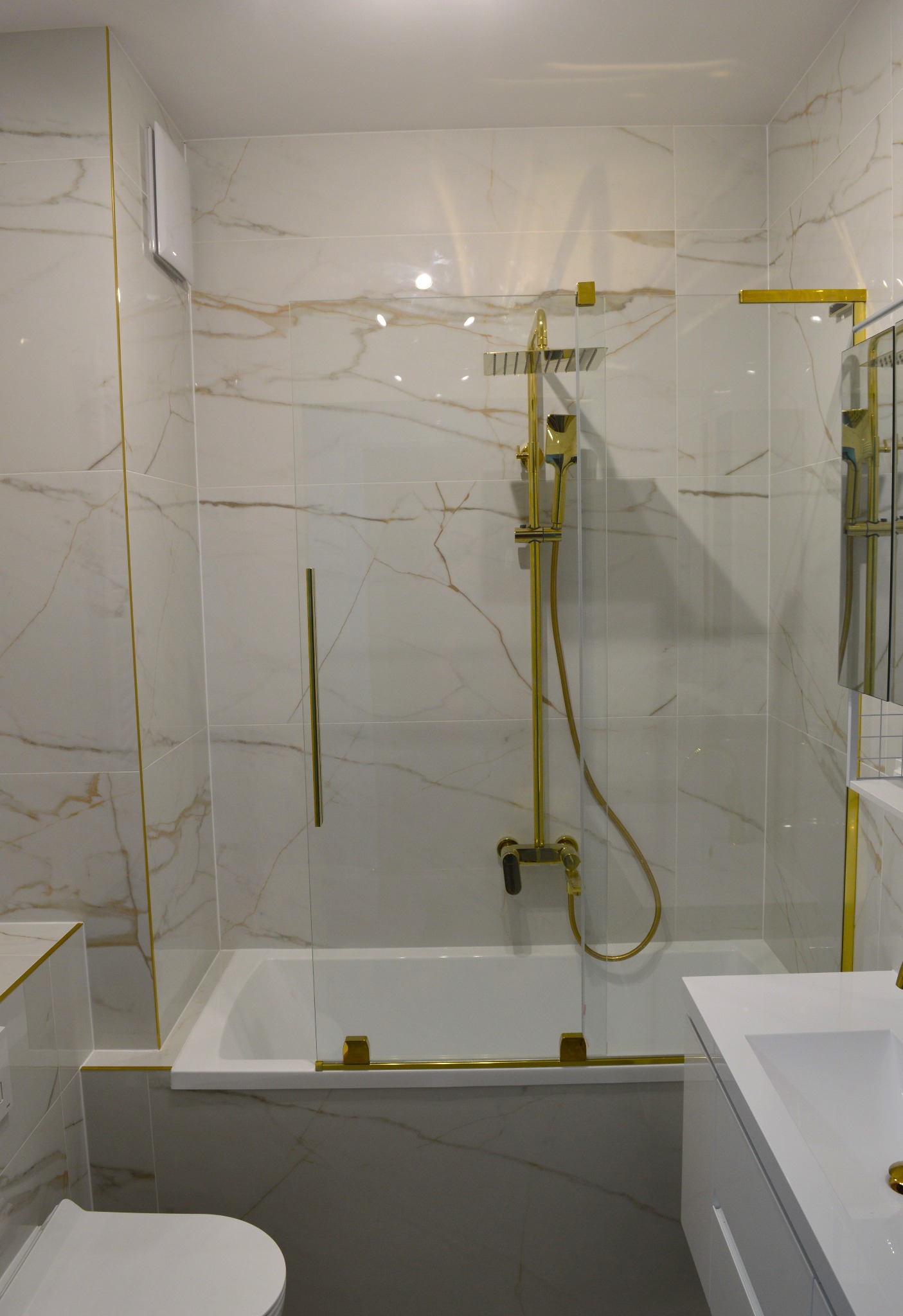 Biała łazienka z marmurowymi płytkami i złotymi akcentami: kabina prysznicowa, bateria, rama lustra. Nowoczesny design z dbałością o detale.