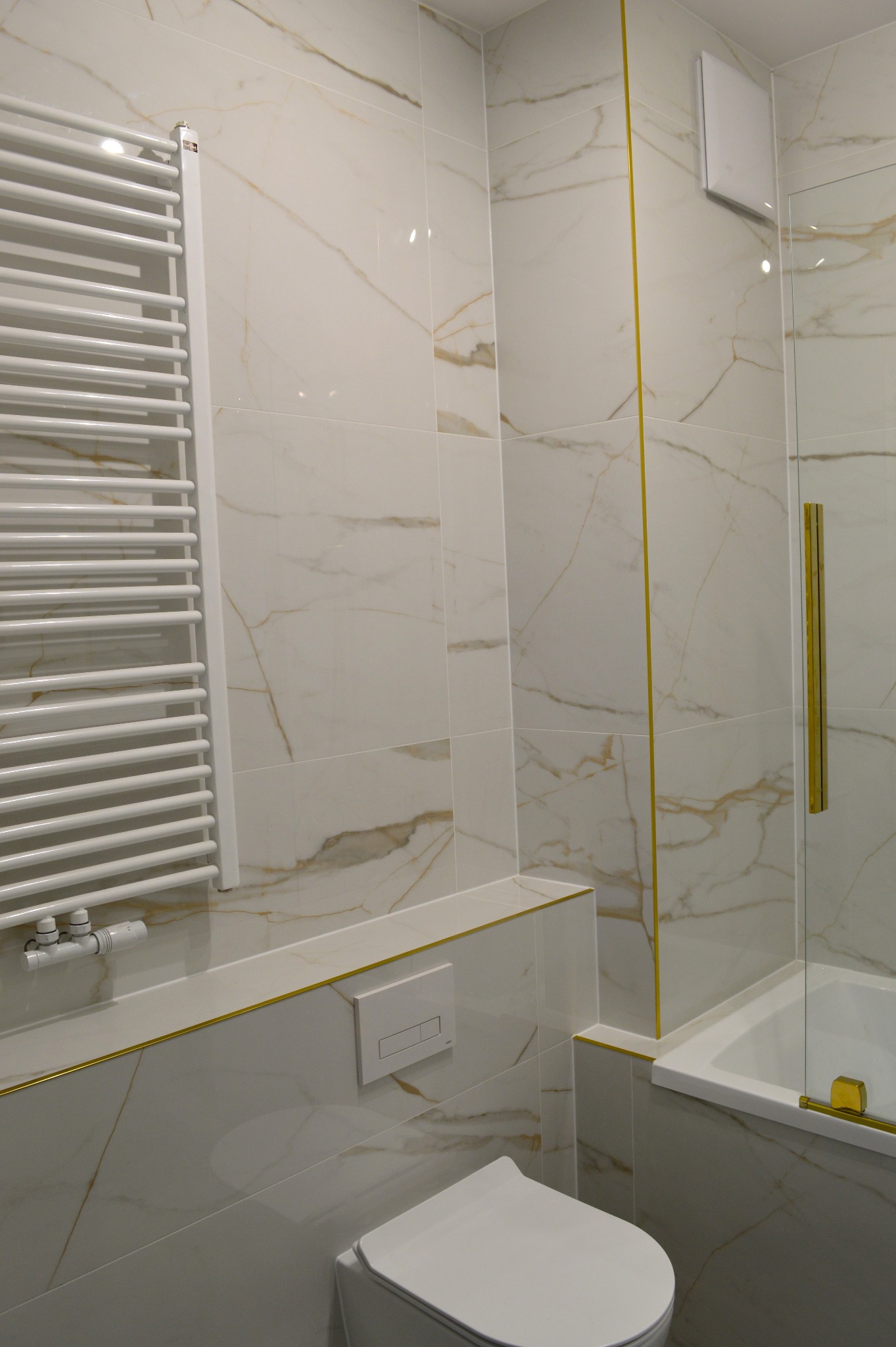 Wykończona łazienka z białymi płytkami imitującymi marmur, złotymi akcentami, grzejnikiem drabinkowym i kabiną prysznicową z uchwytem w kolorze złota.