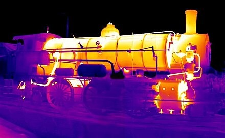 Termowizyjny obraz parowozu, ukazujący rozkład temperatur na jego elementach, z przewagą barw żółtych i fioletowych.