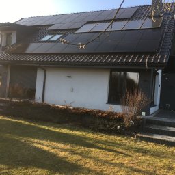 Foto Power Karolina Gajos - Dom jednorodzinny z panelami fotowoltaicznymi na dachu, widoczne okna dachowe i fragment ogrodu z osobą. Ujęcie w słoneczny dzień.