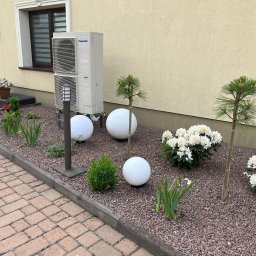 Foto Power Karolina Gajos - Biała jednostka zewnętrzna klimatyzacji Panasonic na tle beżowej elewacji, wkomponowana w rabatę z kulistymi lampami i roślinami ozdobnymi.