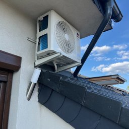 Foto Power Karolina Gajos - Biała klimatyzacja Gree zamontowana na elewacji domu, widoczna z dołu, na tle błękitnego nieba z chmurami. Widoczne elementy dachu i okno.