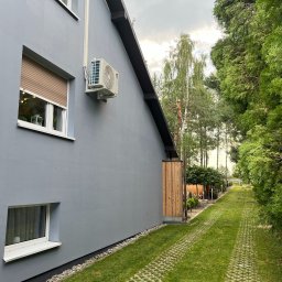 Foto Power Karolina Gajos - Szara elewacja domu z zamontowaną klimatyzacją zewnętrzną, widok z boku na zielony trawnik i drzewa w tle, okno z brązową roletą.