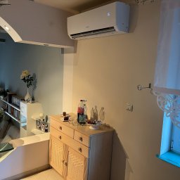 Foto Power Karolina Gajos - Biały klimatyzator GREE zamontowany na ścianie w jasnym salonie. Widoczny fragment okna z firanką i mebel z przekąskami. Nowoczesne, minimalistyczne wnętrze.