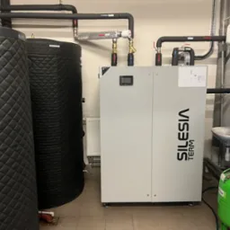 Pompa ciepła Silesia Term w kotłowni z dwoma zasobnikami ciepłej wody, orurowaniem i osprzętem. Widoczna umywalka i zielony zbiornik ciśnieniowy.