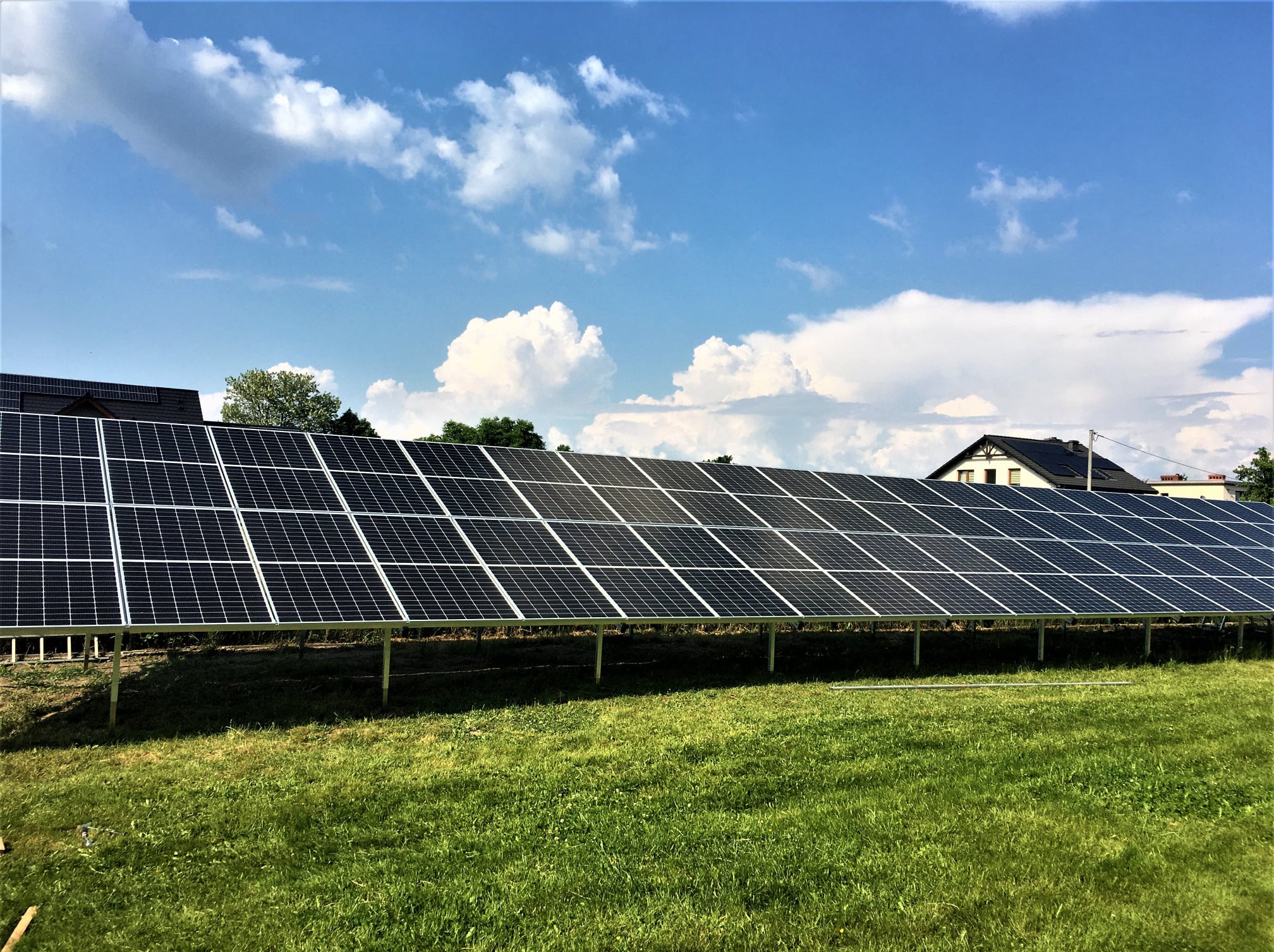 Rząd paneli fotowoltaicznych na metalowej konstrukcji, na tle zielonej trawy, błękitnego nieba z chmurami i zabudowań. Widok z dołu.