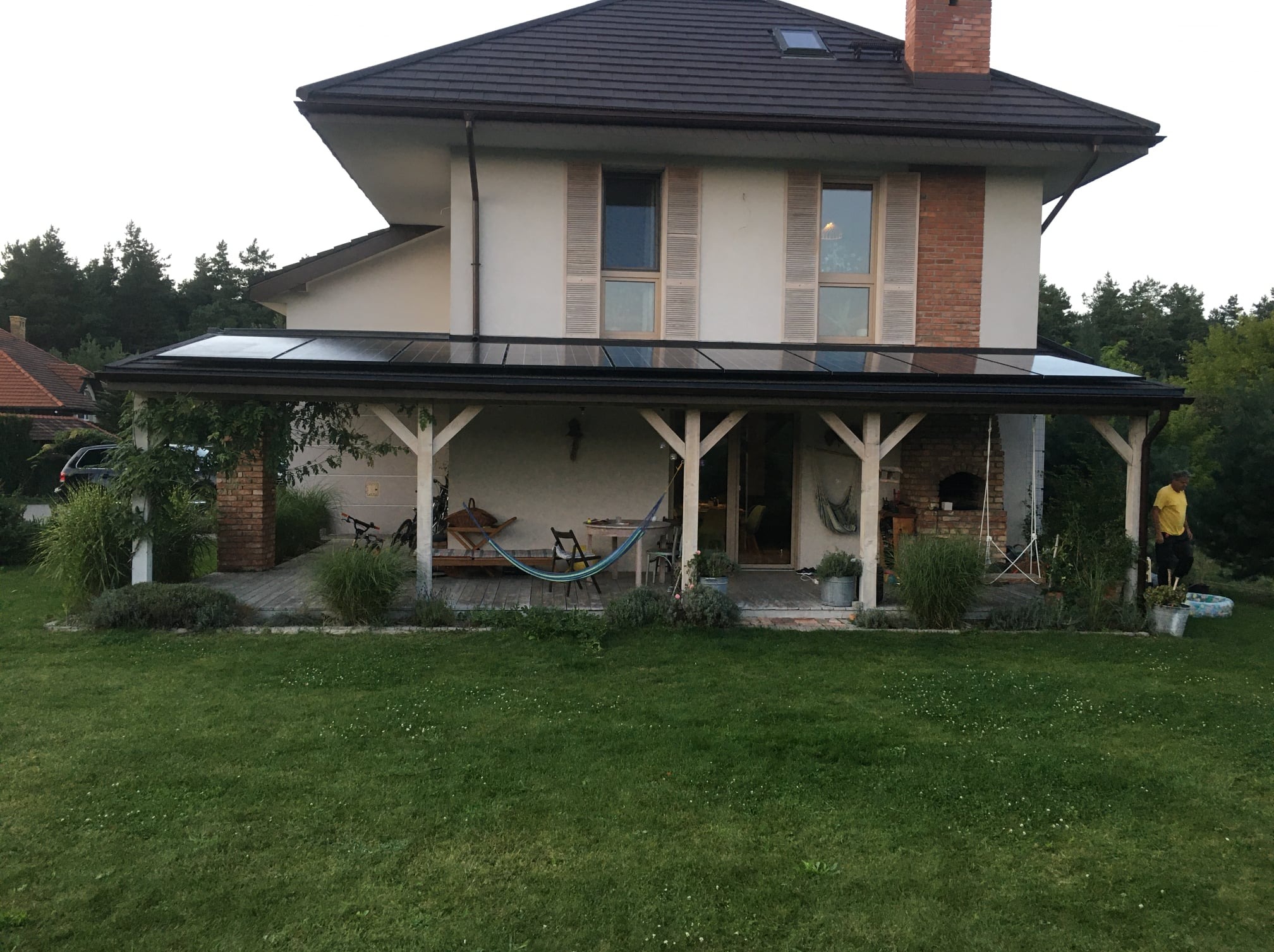 Dom z zadaszonym tarasem, na którym zamontowano panele fotowoltaiczne. Widoczny ogród i osoba w żółtej koszulce.