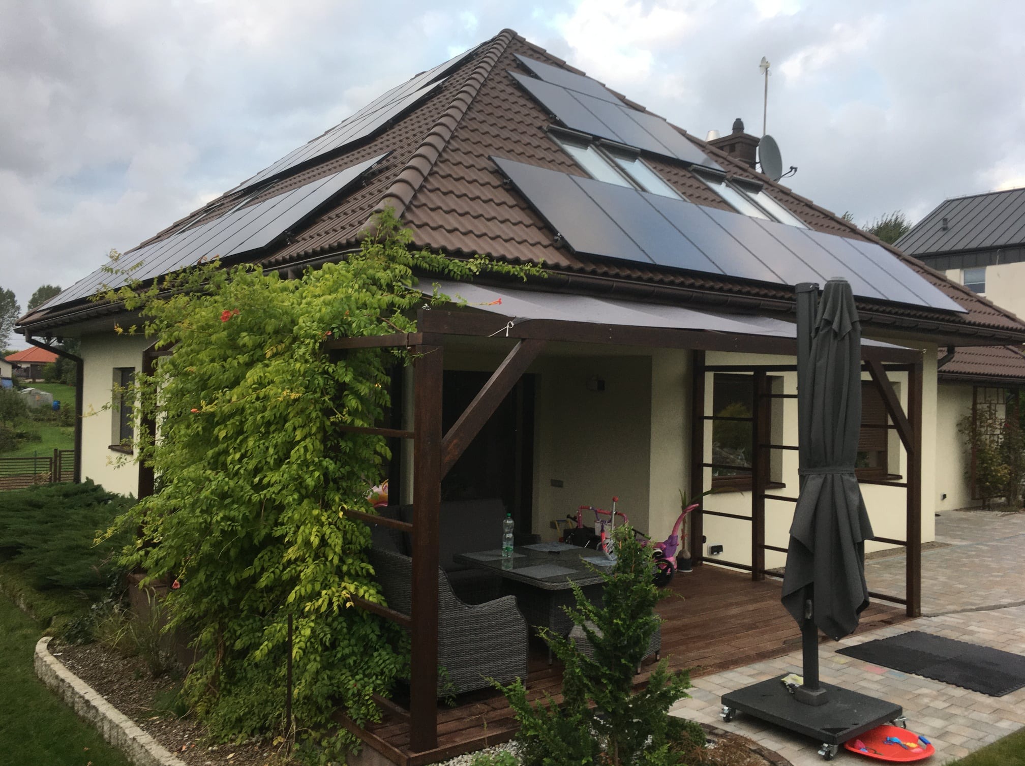 Dom z brązowym dachem i panelami słonecznymi. Widoczny taras z zadaszeniem, rośliny i elementy wyposażenia. Pochmurne niebo w tle.