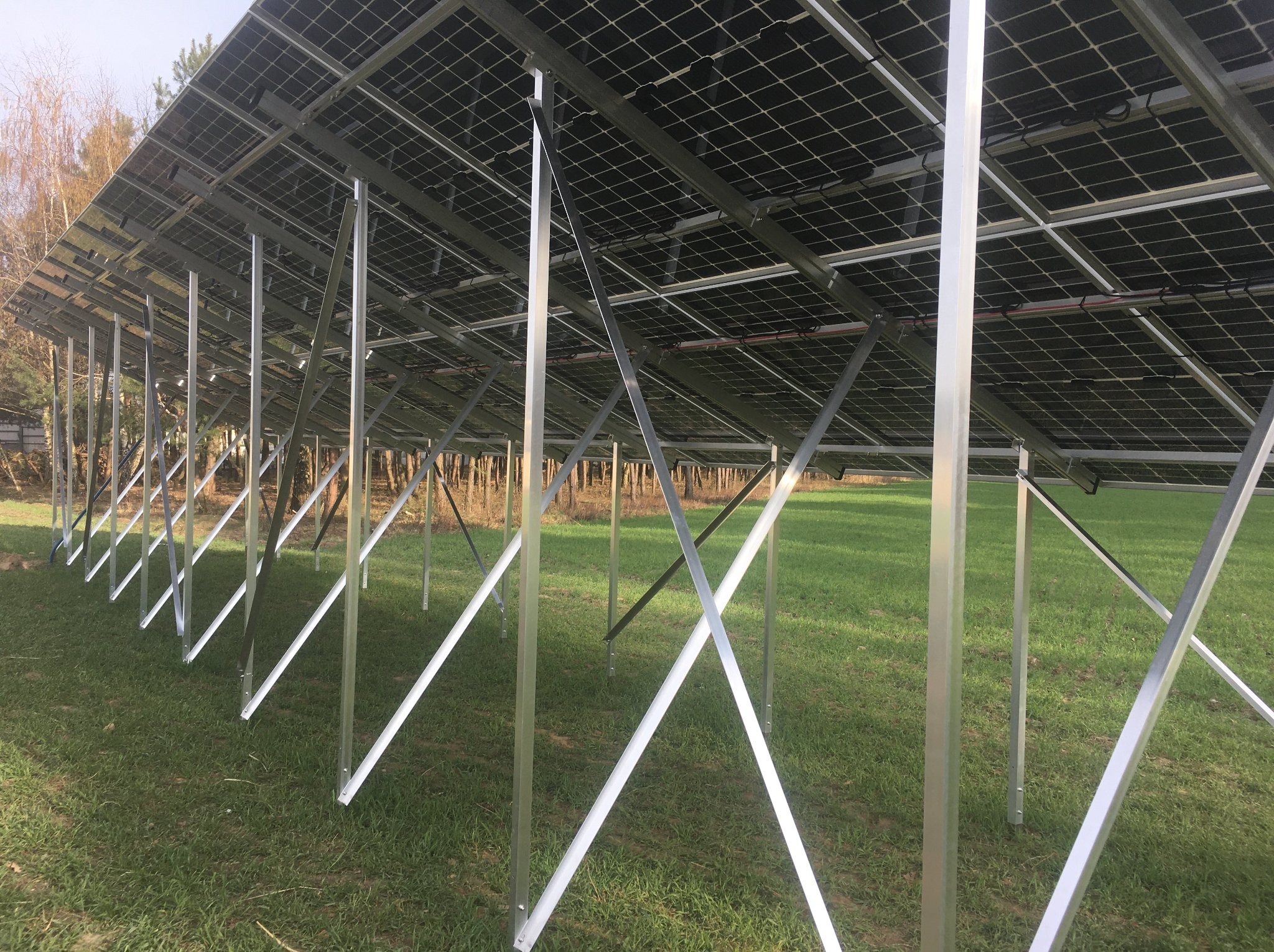 Montaż paneli fotowoltaicznych na metalowej konstrukcji w plenerze. Widok od spodu, ukazujący detale mocowania i rozległe pole z trawą w tle.