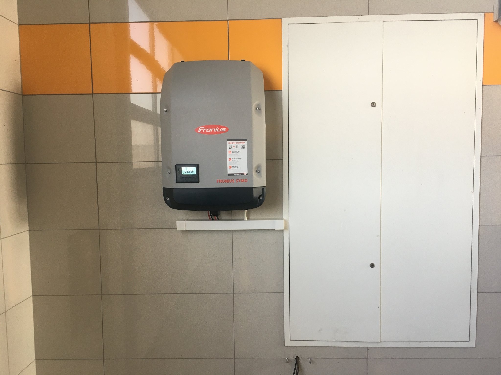 Zamontowany inwerter Fronius na kafelkowanej ścianie, obok biała szafka. Widoczne przewody i panel sterowania. Detal z instalacji fotowoltaicznej w pomieszczeniu.