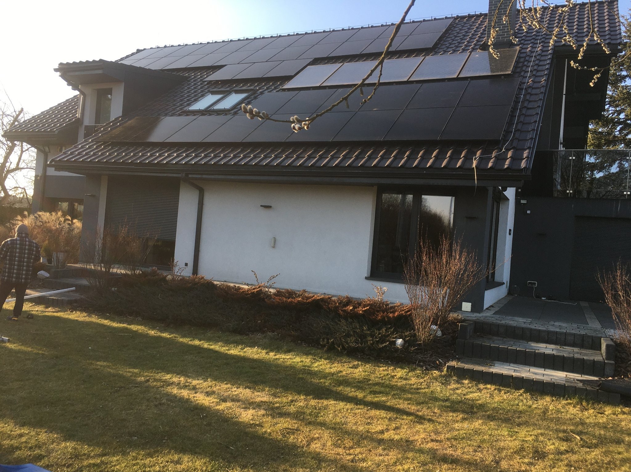 Dom jednorodzinny z panelami fotowoltaicznymi na dachu, widoczne okna dachowe i fragment ogrodu z osobą. Ujęcie w słoneczny dzień.