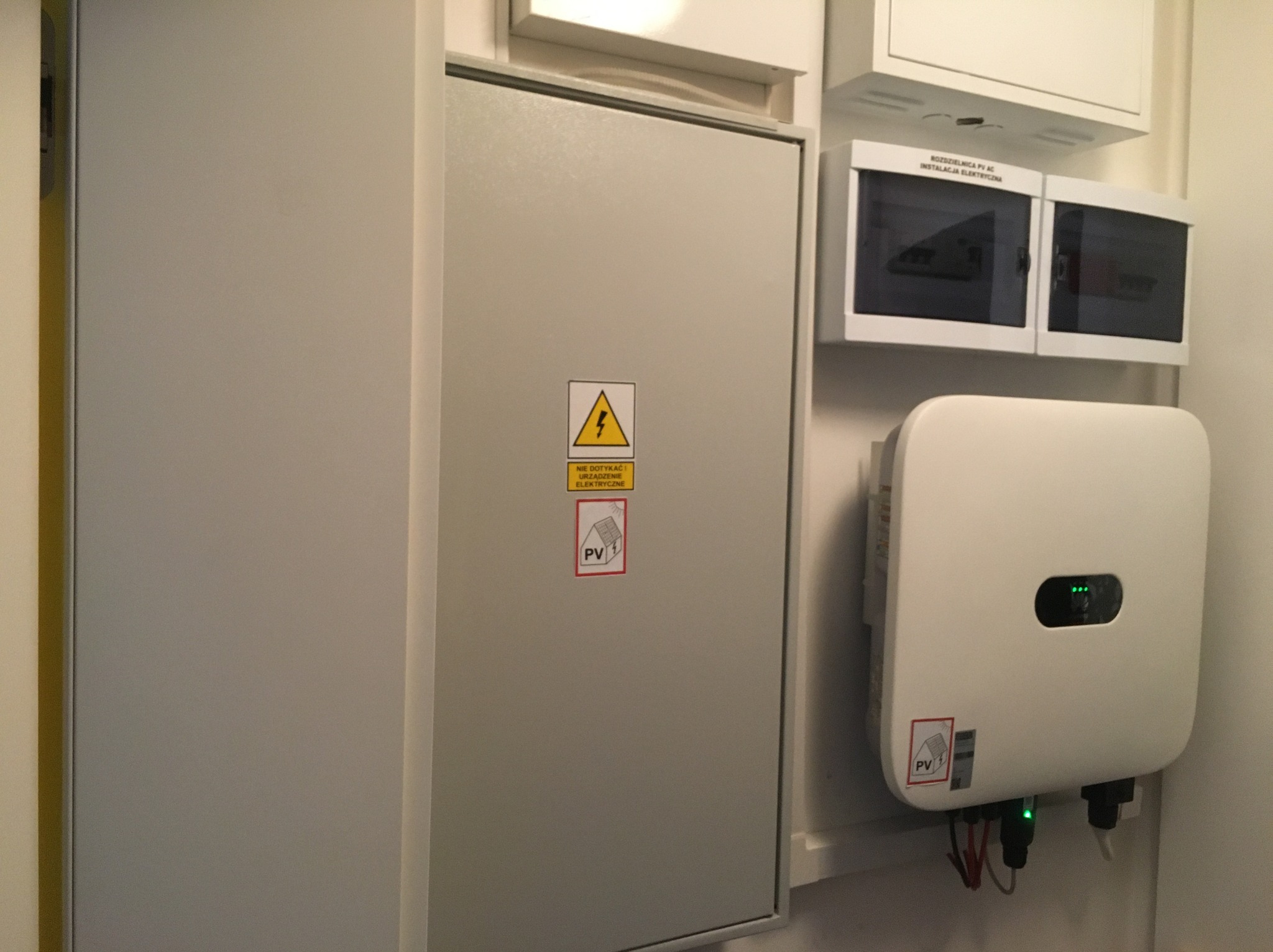 Szara skrzynka elektryczna z oznaczeniem PV i urządzenie z diodami sygnalizującymi pracę instalacji fotowoltaicznej, zamontowane na ścianie wewnątrz budynku.