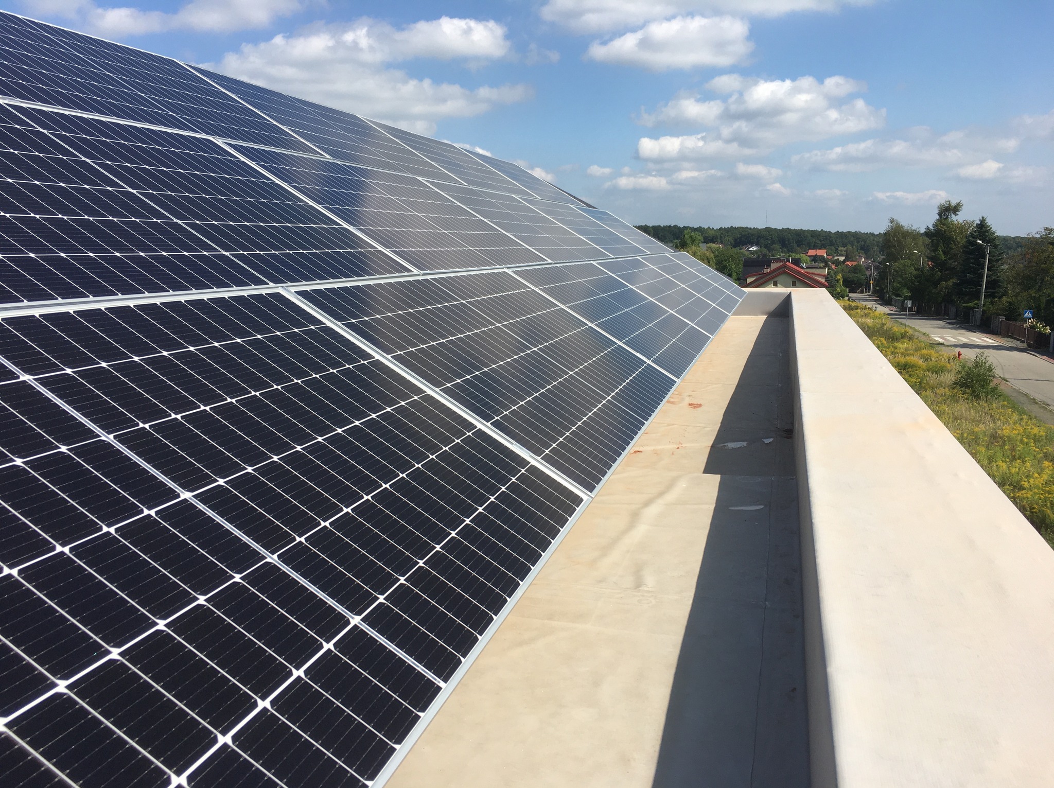 Panele fotowoltaiczne na dachu budynku w słoneczny dzień. Widok z góry, w tle zieleń i zabudowania. Układ paneli w regularnej siatce.
