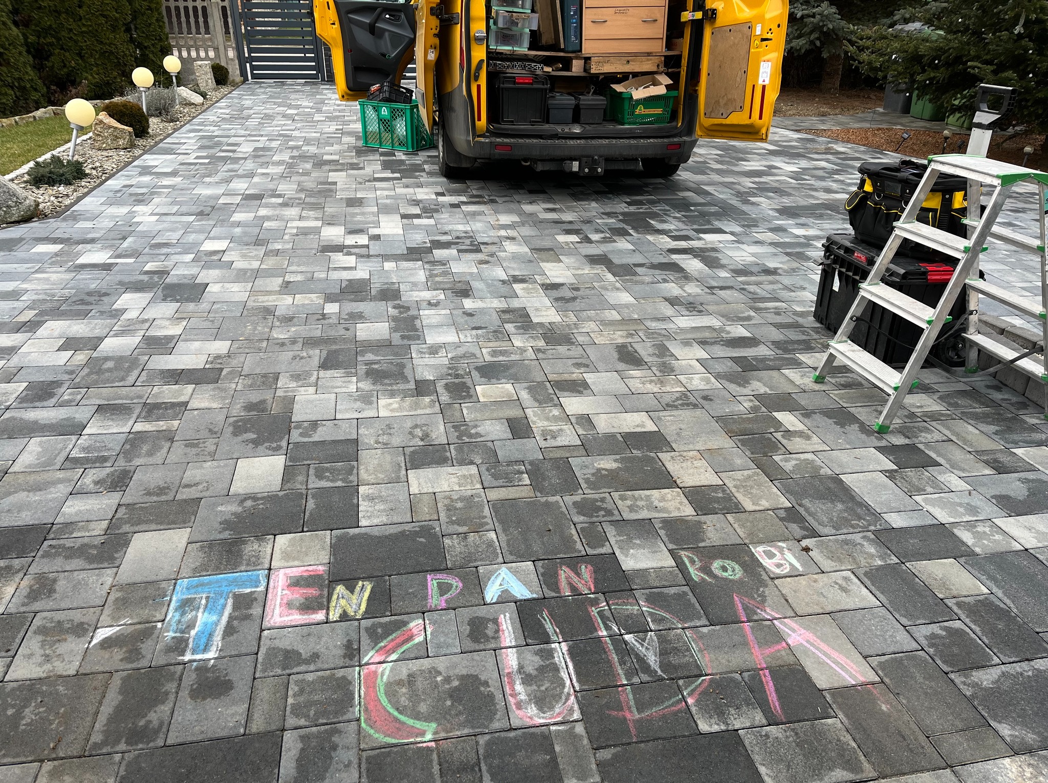 Ułożona kostka brukowa na podjeździe, z napisem kredą 'TEN PAN ROBI CUDA', żółty samochód dostawczy z otwartymi drzwiami, drabina i narzędzia.