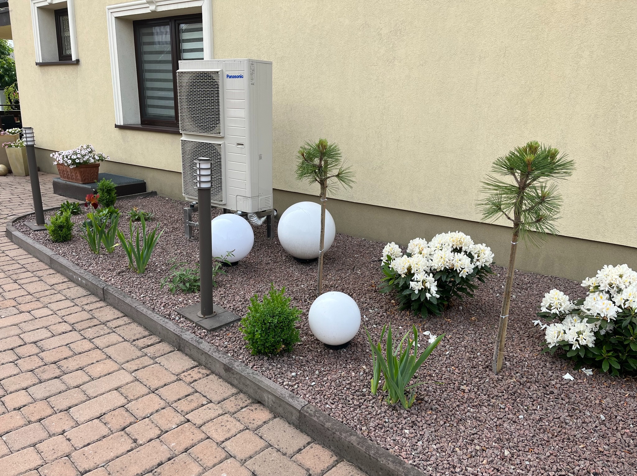 Biała jednostka zewnętrzna klimatyzacji Panasonic na tle beżowej elewacji, wkomponowana w rabatę z kulistymi lampami i roślinami ozdobnymi.