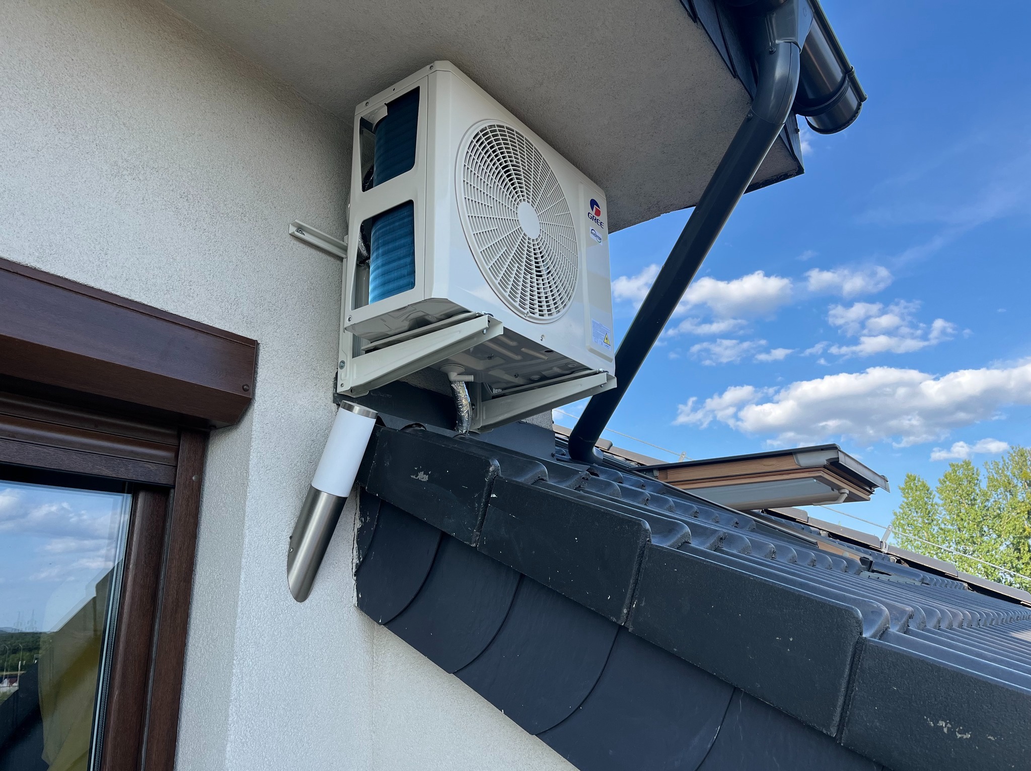 Biała klimatyzacja Gree zamontowana na elewacji domu, widoczna z dołu, na tle błękitnego nieba z chmurami. Widoczne elementy dachu i okno.