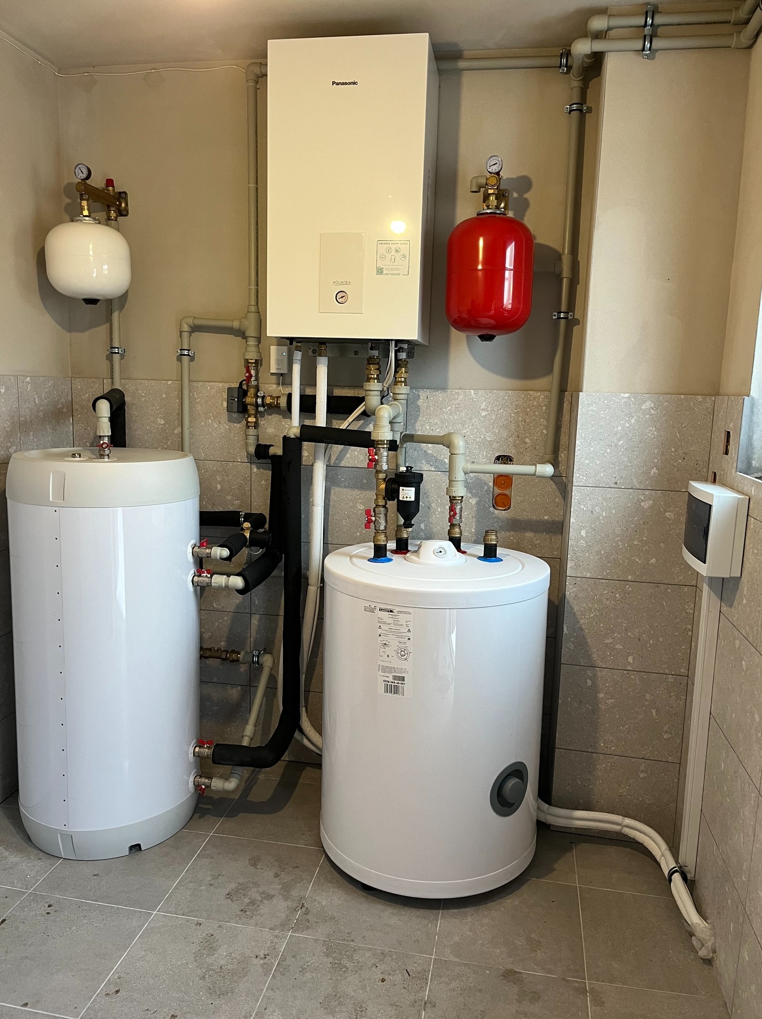 Biały piec gazowy Panasonic z dwoma zbiornikami wody (biały i czerwony) w kotłowni z szarymi płytkami. Widoczne rury, zawory i manometry.