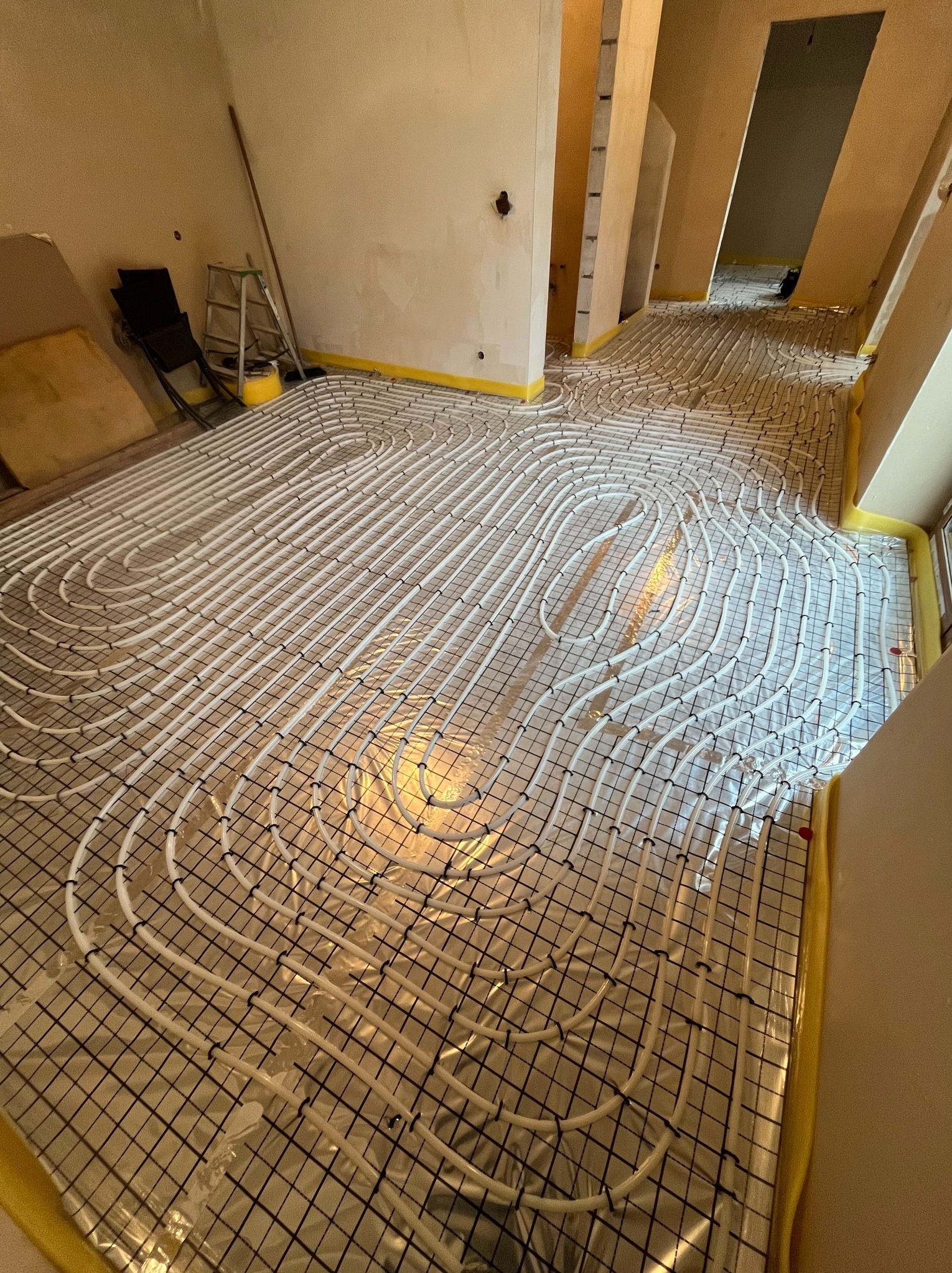 Rozłożona instalacja ogrzewania podłogowego na folii, z widocznymi białymi rurkami i czarną siatką mocującą. Żółta taśma zabezpiecza krawędzie ścian.
