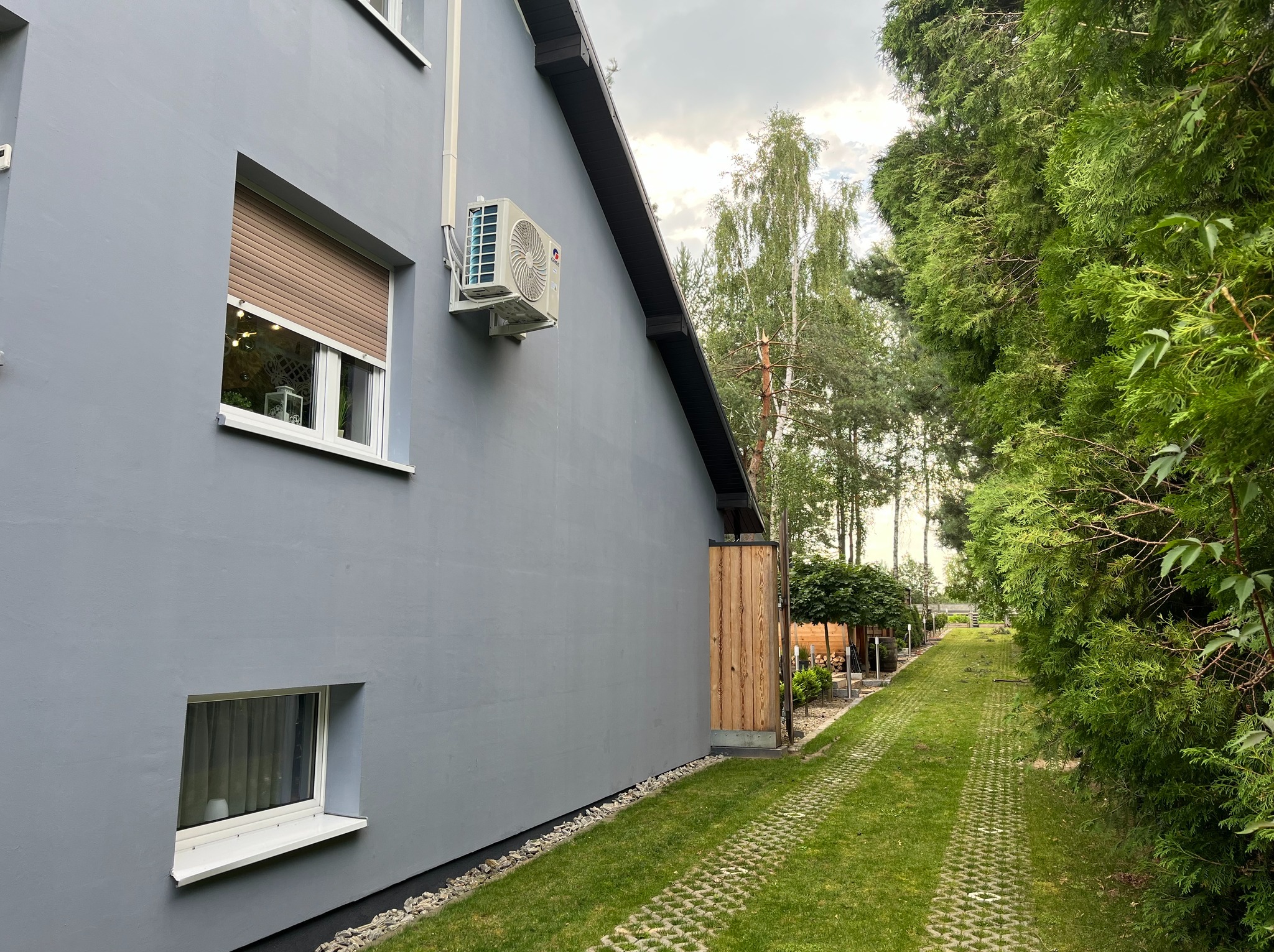 Szara elewacja domu z zamontowaną klimatyzacją zewnętrzną, widok z boku na zielony trawnik i drzewa w tle, okno z brązową roletą.