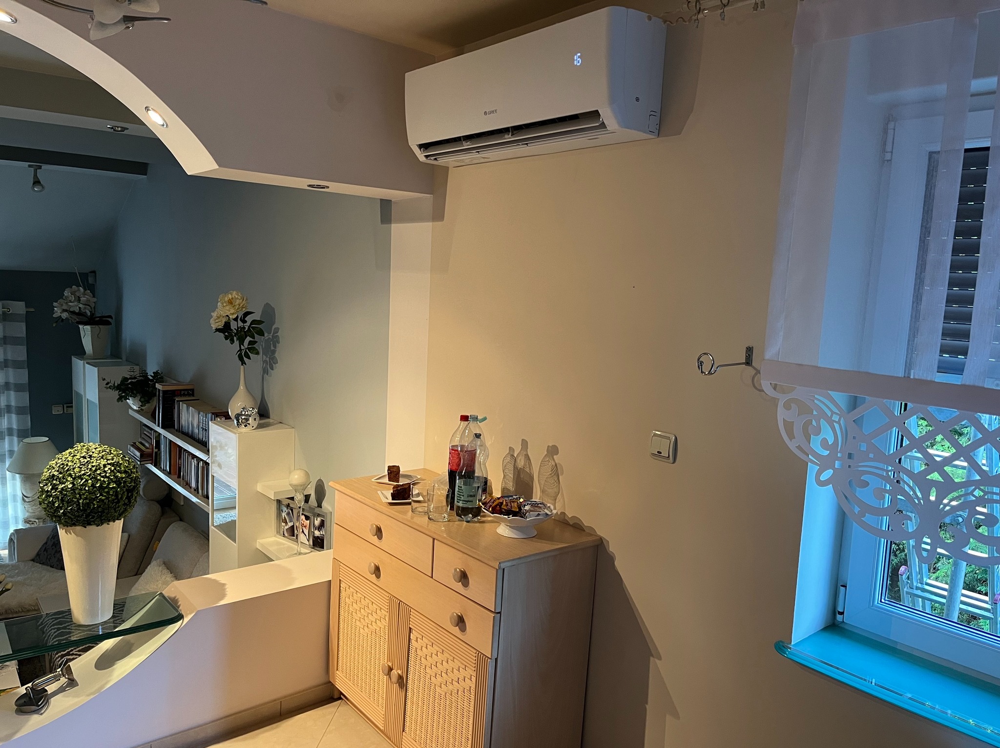 Biały klimatyzator GREE zamontowany na ścianie w jasnym salonie. Widoczny fragment okna z firanką i mebel z przekąskami. Nowoczesne, minimalistyczne wnętrze.