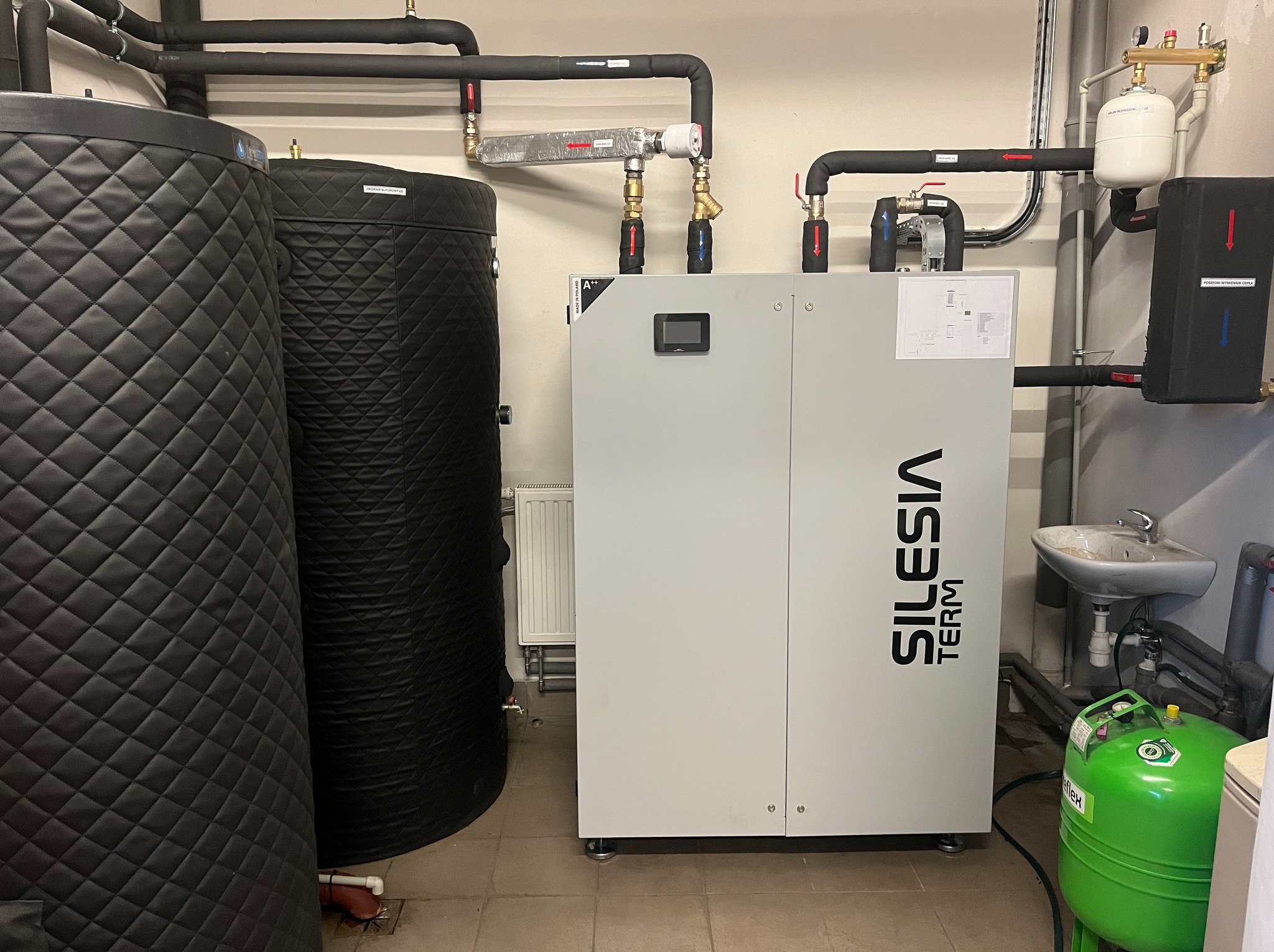 Pompa ciepła Silesia Term w kotłowni z dwoma zasobnikami ciepłej wody, orurowaniem i osprzętem. Widoczna umywalka i zielony zbiornik ciśnieniowy.