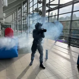 Osoba w kombinezonie taktycznym strzela dymem z karabinu podczas imprezy laser tag w hali z dmuchanymi przeszkodami.