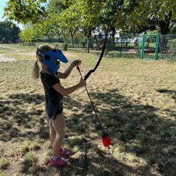 TOSA SPÓŁKA Z OGRANICZONĄ ODPOWIEDZIALNOŚCIĄ - Dziewczynka w masce celuje z łuku na trawiastym terenie, przygotowując się do strzału. W tle widoczne drzewa i ogrodzenie.