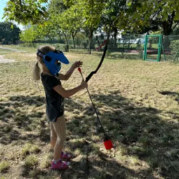 Dziewczynka w masce celuje z łuku na trawiastym terenie, przygotowując się do strzału. W tle widoczne drzewa i ogrodzenie.