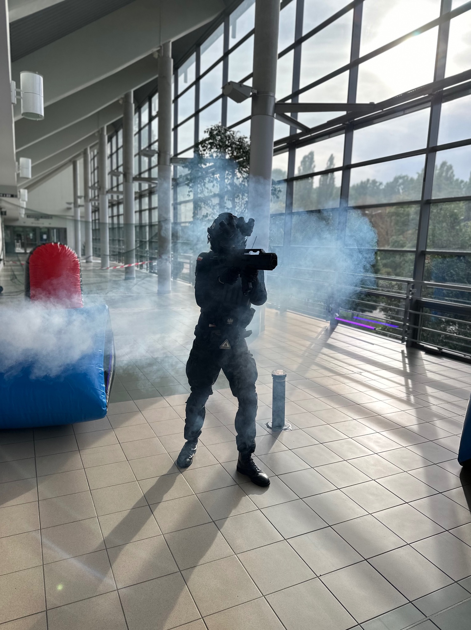 Osoba w kombinezonie taktycznym strzela dymem z karabinu podczas imprezy laser tag w hali z dmuchanymi przeszkodami.