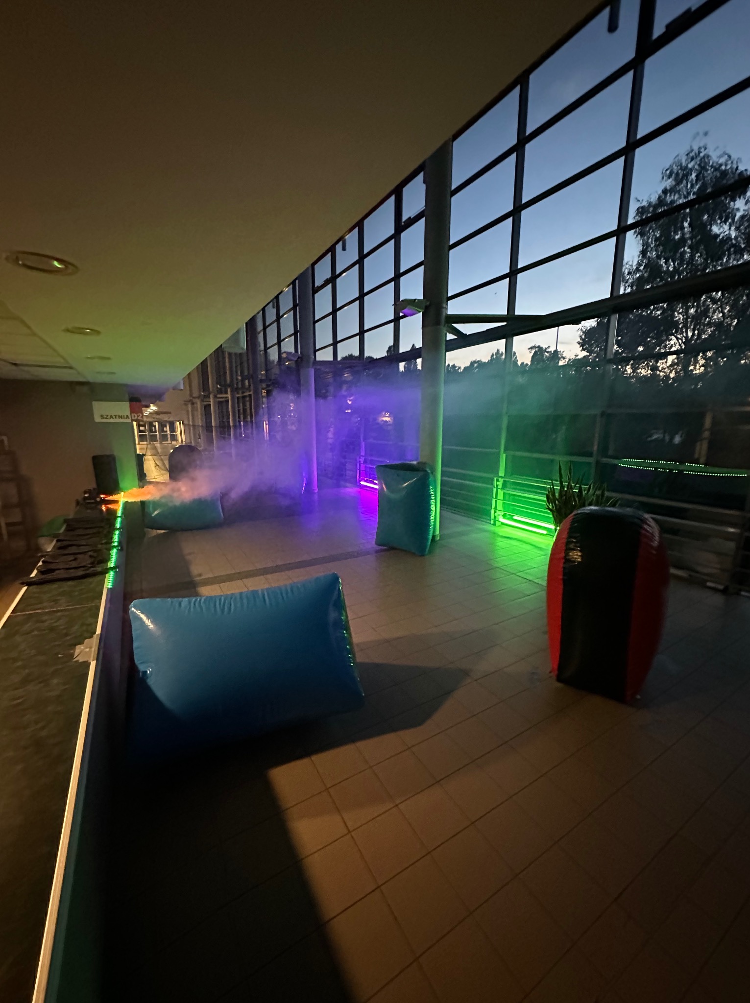 Przygotowane pole do laserowego paintballa z dmuchanymi przeszkodami, oświetlone kolorowymi światłami i wypełnione dymem, gotowe na imprezę integracyjną.