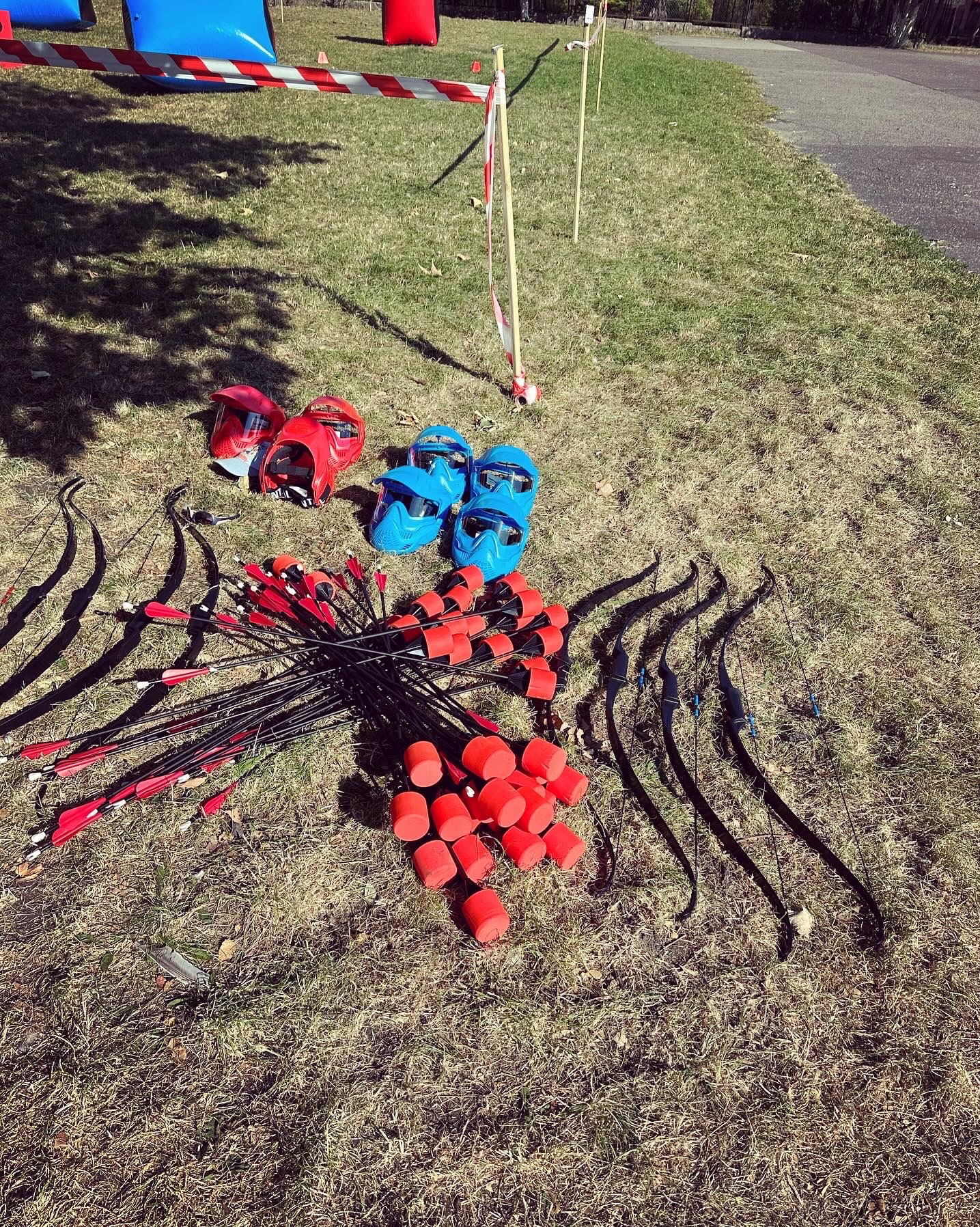 Łuki, strzały z miękkimi końcówkami i kaski ochronne na trawie, przygotowane do archery tag. Taśmy wyznaczają teren gry.