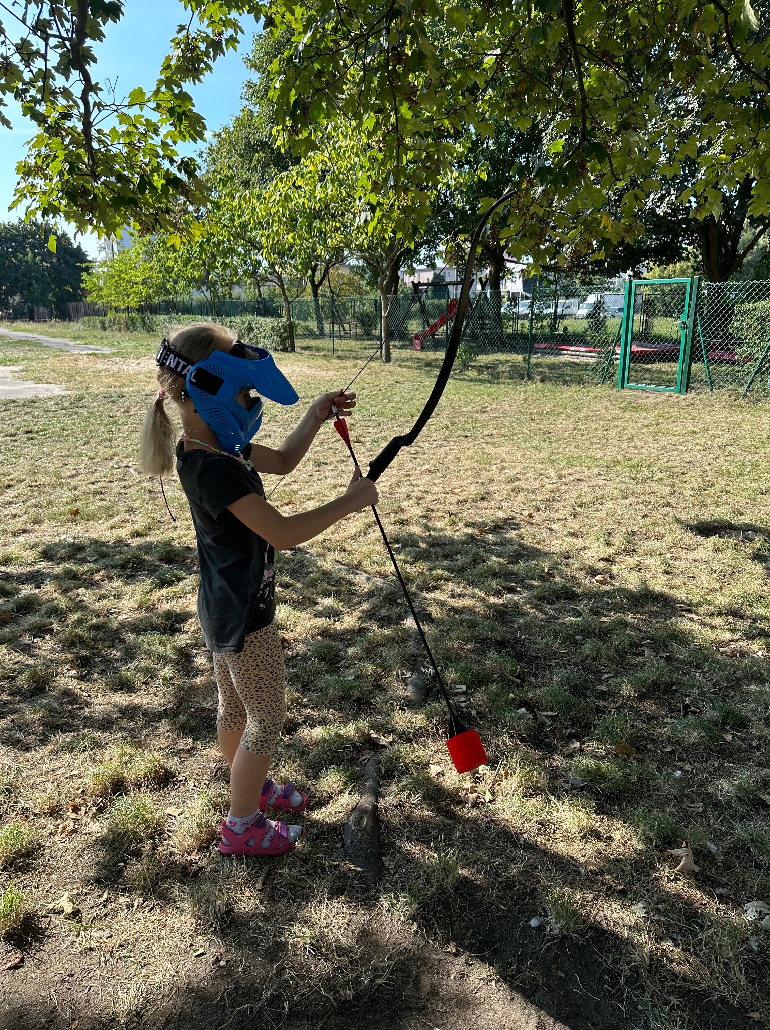 Dziewczynka w masce celuje z łuku na trawiastym terenie, przygotowując się do strzału. W tle widoczne drzewa i ogrodzenie.