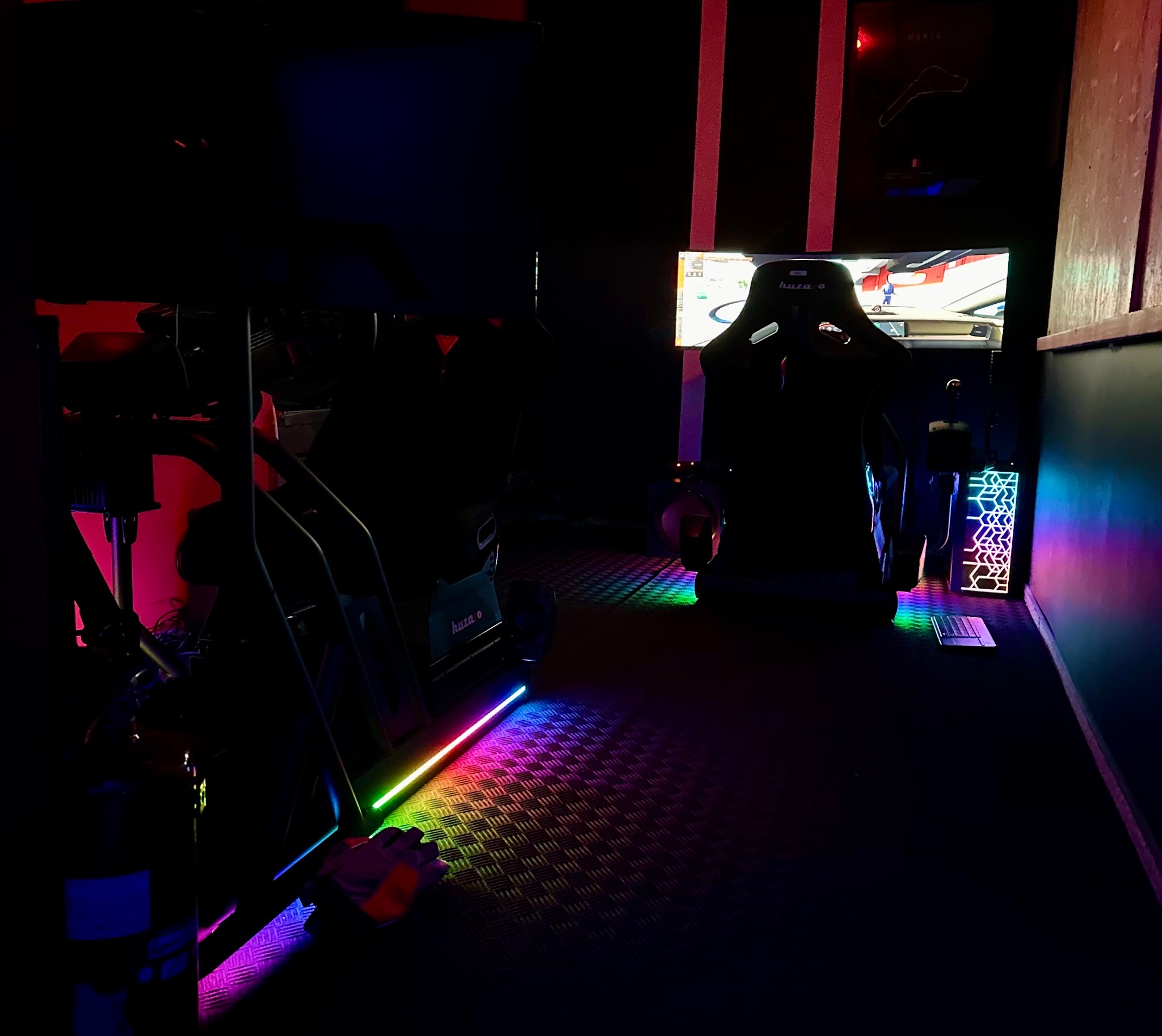 Dwa stanowiska symulatorów wyścigowych w ciemnym pomieszczeniu, podświetlone kolorowymi ledami, z ekranami wyświetlającymi grę. Klimatyczna przestrzeń do rozrywki.