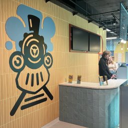 MANAGEMENT GROUP SPÓŁKA Z OGRANICZONĄ ODPOWIEDZIALNOŚCIĄ - Recepcja z dekoracyjną grafiką lokomotywy na ścianie z beżowych płytek. Za ladą widoczni klienci.