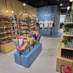 MANAGEMENT GROUP SPÓŁKA Z OGRANICZONĄ ODPOWIEDZIALNOŚCIĄ - Wnętrze sklepu z pamiątkami: półki z zabawkami, poduszkami i koszulkami. Jasne oświetlenie, kolorowe wyposażenie, osoba w tle. Ujęcie z perspektywy klienta.