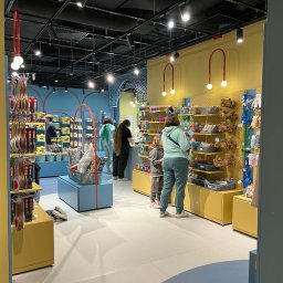 MANAGEMENT GROUP SPÓŁKA Z OGRANICZONĄ ODPOWIEDZIALNOŚCIĄ - Wnętrze sklepu z zabawkami i akcesoriami dla dzieci, z żółto-niebieską kolorystyką, oświetleniem punktowym i klientami przy regałach.