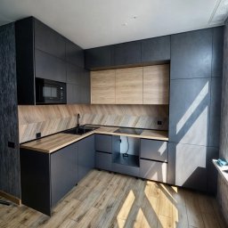 MANAGEMENT GROUP SPÓŁKA Z OGRANICZONĄ ODPOWIEDZIALNOŚCIĄ - Nowoczesna kuchnia z ciemnymi, matowymi szafkami i drewnianymi akcentami, oświetlona promieniami słońca padającymi na drewnianą podłogę.