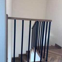 MANAGEMENT GROUP SPÓŁKA Z OGRANICZONĄ ODPOWIEDZIALNOŚCIĄ - Nowoczesne schody z drewnianymi stopniami i czarną, metalową balustradą z drewnianym poręczą, widok z góry, parkiet ułożony w jodełkę.