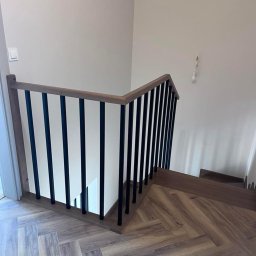 MANAGEMENT GROUP SPÓŁKA Z OGRANICZONĄ ODPOWIEDZIALNOŚCIĄ - Nowoczesne schody z drewnianymi stopniami i czarną, metalową balustradą. Parkiet ułożony w jodełkę. Minimalistyczny design.