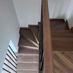 MANAGEMENT GROUP SPÓŁKA Z OGRANICZONĄ ODPOWIEDZIALNOŚCIĄ - Drewniane schody z czarną, metalową balustradą, widok z góry. Stopnie wykończone parkietem w jodełkę, kontrastujące odcienie drewna. Minimalistyczny design.