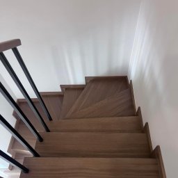 MANAGEMENT GROUP SPÓŁKA Z OGRANICZONĄ ODPOWIEDZIALNOŚCIĄ - Kątowe ujęcie drewnianych schodów z czarną, minimalistyczną balustradą. Widok z góry podkreśla geometrię stopni i kontrast materiałów.