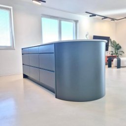 MANAGEMENT GROUP SPÓŁKA Z OGRANICZONĄ ODPOWIEDZIALNOŚCIĄ - Nowoczesna wyspa kuchenna z szufladami w odcieniach szarości, zaokrąglony narożnik, blat z jasnego kamienia, minimalistyczne oświetlenie, jasna podłoga.