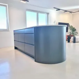 Nowoczesna wyspa kuchenna z szufladami w odcieniach szarości, zaokrąglony narożnik, blat z jasnego kamienia, minimalistyczne oświetlenie, jasna podłoga.