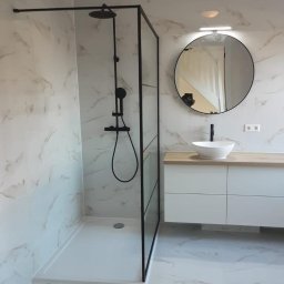 MANAGEMENT GROUP SPÓŁKA Z OGRANICZONĄ ODPOWIEDZIALNOŚCIĄ - Nowoczesna łazienka z kabiną typu walk-in, czarną armaturą i okrągłym lustrem. Marmurowe płytki na ścianach i podłodze, biała szafka pod umywalką z drewnianym blatem.